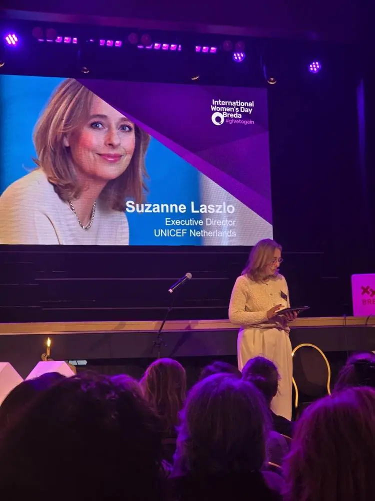 A presenter on stage beside a screen showing the name and title of the guest speaker