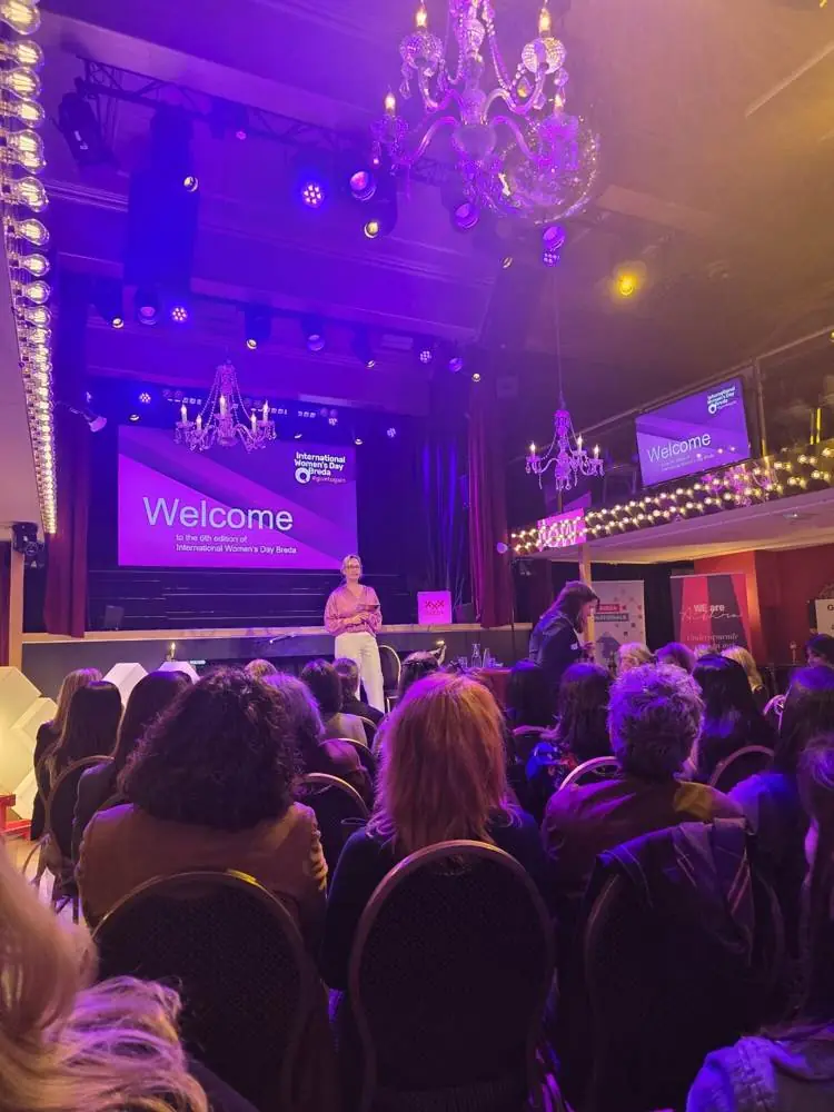 A speaker on stage at the International Women's Day event, with a welcome screen behind her